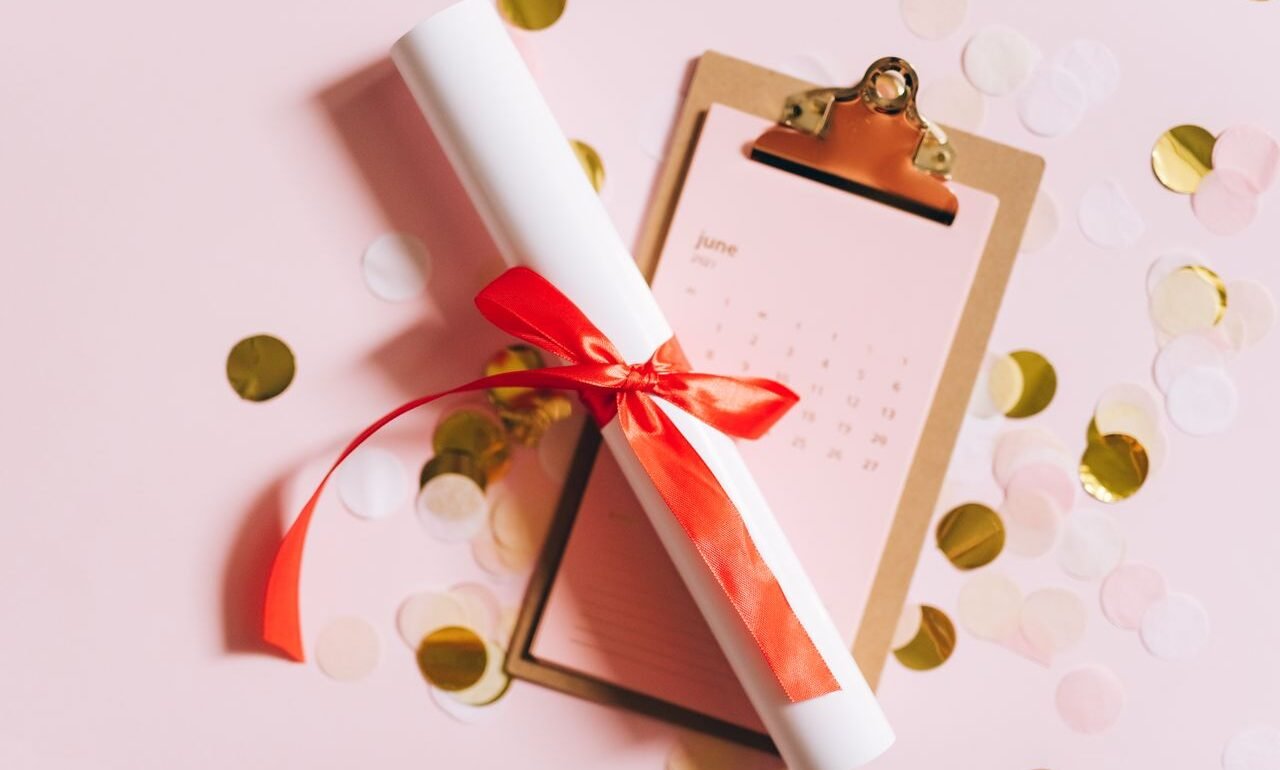 Flat lay of a graduation diploma with a red ribbon on a clipboard with confetti.